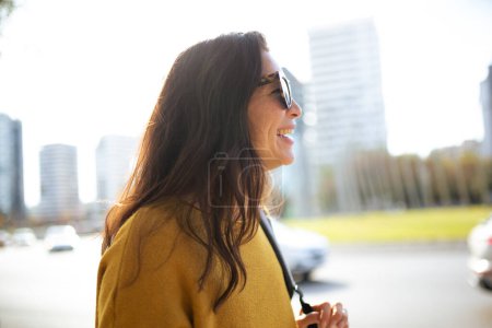Téléchargez les photos : Portrait latéral de jolie jeune femme avec des lunettes de soleil marchant à l'extérieur dans la ville - en image libre de droit