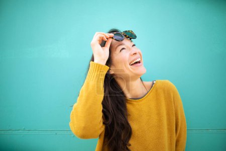 Foto de Primer plano retrato de alegre joven asiática mujer con gafas de sol mirando hacia otro lado y sonriendo contra la pared verde - Imagen libre de derechos