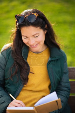 Foto de Retrato de cerca de una joven feliz sentada en un banco escribiendo en un libro afuera - Imagen libre de derechos