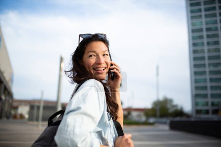 Photo for Rear view portrait of young asian woman talking on mobile phone and looking behind while walking outside in city - Royalty Free Image