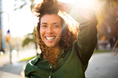 Téléchargez les photos : Gros plan portrait de belle jeune femme latine regardant loin et souriant à l'extérieur avec le soleil qui brille par derrière - en image libre de droit
