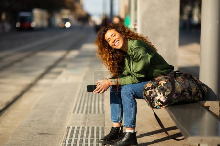 Téléchargez les photos : Portrait de jeune voyageuse séduisante avec téléphone en attente à la gare de tramway en ville - en image libre de droit