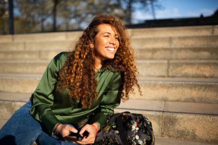 Téléchargez les photos : Portrait de belle femme hispanique assise sur des marches à l'extérieur avec téléphone portable et sac regardant loin et souriant - en image libre de droit
