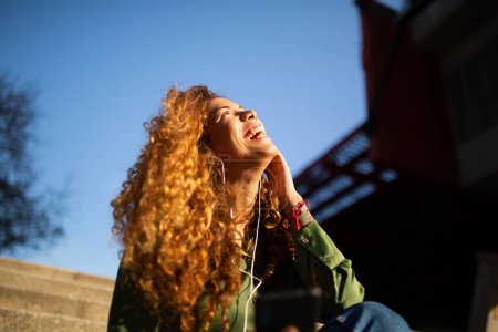 Foto de Retrato de una joven sonriente sentada afuera en la ciudad con auriculares disfrutando escuchando música - Imagen libre de derechos