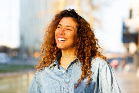 Téléchargez les photos : Gros plan portrait de jolie jeune femme hispanique regardant loin et souriant debout dehors dans la ville - en image libre de droit