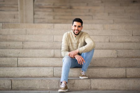 Photo for Full length portrait of handsome young arab man sitting on steps looking at camera and smiling - Royalty Free Image