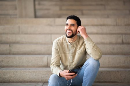Téléchargez les photos : Portrait de jeune homme arabe heureux écoutant de la musique avec un téléphone portable assis sur les marches - en image libre de droit