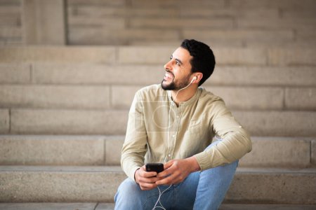 Photo for Portrait of smiling arabic man listening to music with mobile phone sitting on steps - Royalty Free Image