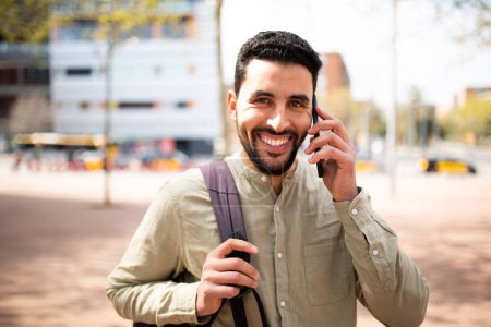 Téléchargez les photos : Portrait d'un jeune homme arabe souriant parlant sur un téléphone portable marchant dehors dans la ville - en image libre de droit
