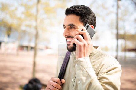 Téléchargez les photos : Portrait latéral d'un beau jeune homme arabe marchant dehors parlant sur un téléphone portable - en image libre de droit