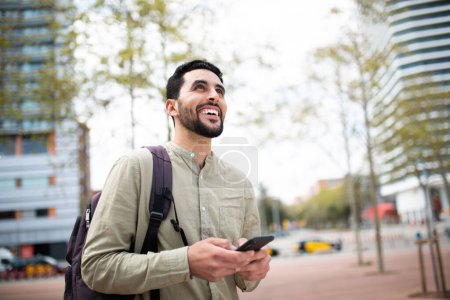 Téléchargez les photos : Portrait de jeune homme heureux marchant dehors dans la ville avec sac et téléphone portable - en image libre de droit