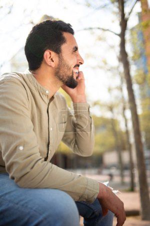 Téléchargez les photos : Portrait latéral d'un jeune homme arabe souriant parlant sur un téléphone portable assis à l'extérieur - en image libre de droit