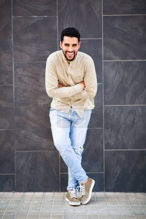 Téléchargez les photos : Portrait complet du jeune homme arabe souriant debout contre un mur noir avec les bras croisés regardant la caméra - en image libre de droit