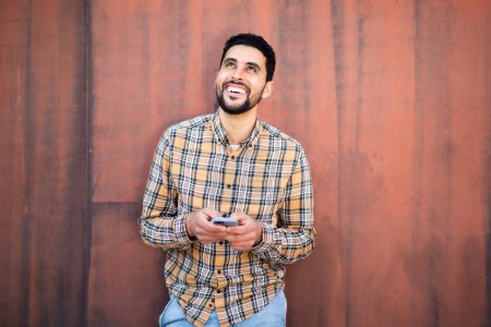 Photo for Portrait of handsome young arabic man standing with cell phone against brown wall looking away and smiling - Royalty Free Image