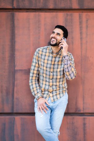 Foto de Retrato de un hombre árabe joven y elegante hablando en el teléfono móvil contra la pared marrón mirando hacia otro lado y sonriendo - Imagen libre de derechos