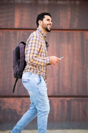 Photo for Side portrait of cheerful young arabic guy walking outside with bag and mobile phone - Royalty Free Image