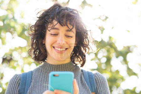 Téléchargez les photos : Gros plan portrait de jolie jeune femme regardant un téléphone portable debout dehors par une journée ensoleillée - en image libre de droit