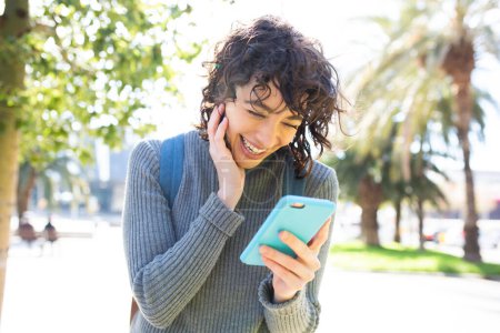 Photo for Close-up portrait of pretty young woman looking at cellphone and smiling while walking outside in the city - Royalty Free Image