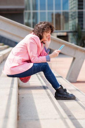Photo for Side portrait of happy young woman sitting on steps in city with mobile phone - Royalty Free Image