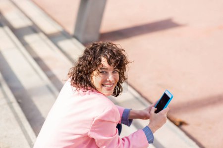 Photo for Rear view portrait of pretty young woman with mobile phone looking behind and smiling while sitting on steps - Royalty Free Image