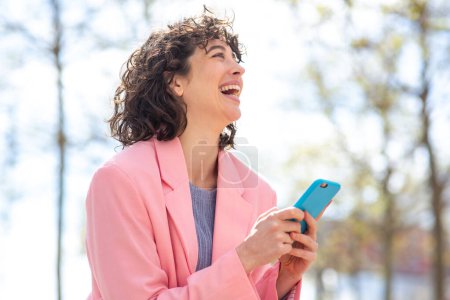 Foto de Retrato de una joven alegre con teléfono mirando hacia otro lado y riendo de pie al aire libre - Imagen libre de derechos