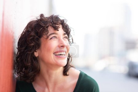 Foto de Primer plano retrato de la joven alegre inclinada a la pared exterior mirando hacia otro lado y sonriendo - Imagen libre de derechos