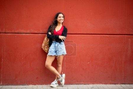 Foto de Retrato de cuerpo completo de una hermosa joven de pie junto a la pared roja con los brazos cruzados - Imagen libre de derechos