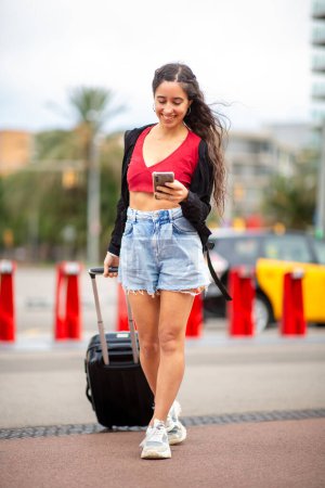 Téléchargez les photos : Portrait corporel complet d'une jeune voyageuse marchant avec valise et téléphone portable - en image libre de droit