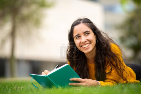 Téléchargez les photos : Portrait de jeune femme souriante allongée sur l'herbe avec un livre et un stylo - en image libre de droit