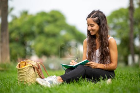 Téléchargez les photos : Portrait de jeune femme assise sur l'herbe et écrivant dans un livre - en image libre de droit
