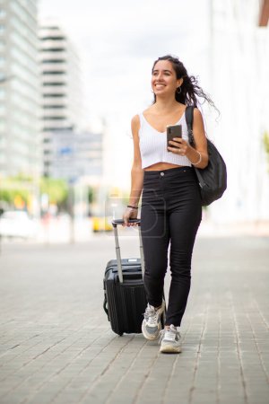 Téléchargez les photos : Portrait complet de jeune voyageuse marchant avec téléphone portable et valise - en image libre de droit