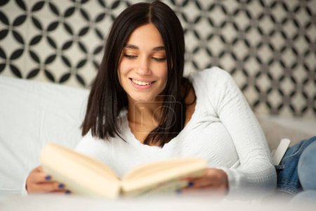 Foto de Retrato sonriente joven mujer relajante en la cama libro de lectura - Imagen libre de derechos
