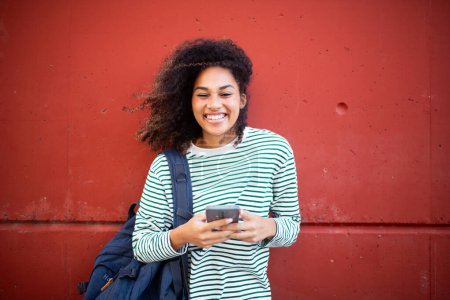 Téléchargez les photos : Portrait heureuse jeune femme afro-américaine debout au mur avec téléphone portable et sac - en image libre de droit