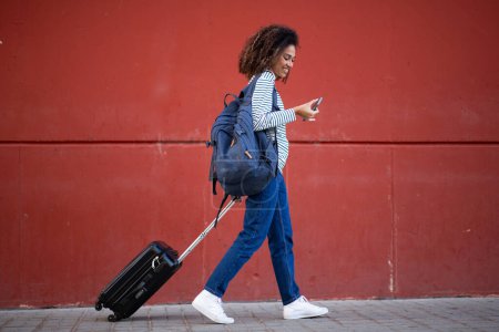 Foto de Retrato de cuerpo completo joven mujer de viaje tirando de la maleta y mirando el teléfono - Imagen libre de derechos