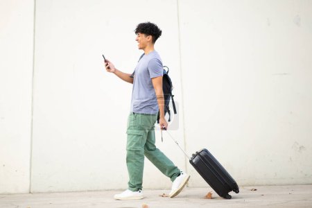 Foto de Retrato lateral de cuerpo completo joven caminando con teléfono móvil y bolsa de viaje - Imagen libre de derechos
