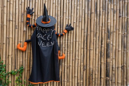 Téléchargez les photos : Arrière de sorcier avec chapeau noir et peinture manteau Joyeux Halloween attaché sur le mur de bambou. Décoration extérieure de vacances. - en image libre de droit