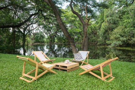 Détendez-vous transat en bois et table sur pelouse verte cour par étang avec de beaux grands arbres et réflexion sur l'eau. Café extérieur et restaurant.