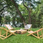 Relax wooden deckchair and table on green grass yard by pond with beautiful big trees and reflection on water. Outdoor cafe and restaurant.