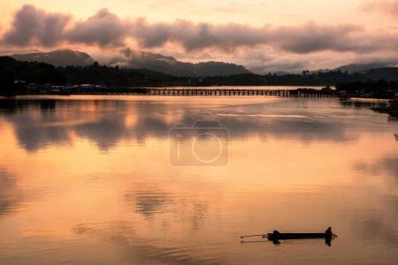 Silueta pescador pescado en barco por la madera Mon puente con reflejo de luz al amanecer en el río Songkalia, Sangkhlaburi, Kanchanaburi, Tailandia. Famoso destino turístico o turista en Siam