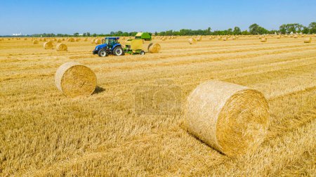 Photo for Above view round bale of straw after harvest, in background tractor is pulling round baler, machine that rolls up the straw and spits out a packed them over agricultural field. - Royalty Free Image