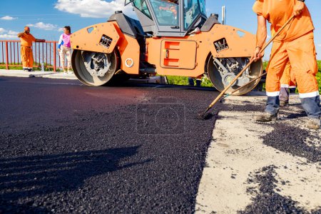 Photo for Workers are using rakes to level, set up layer of fresh tarmac to right measures, steamroller is flatting fresh asphalt . - Royalty Free Image