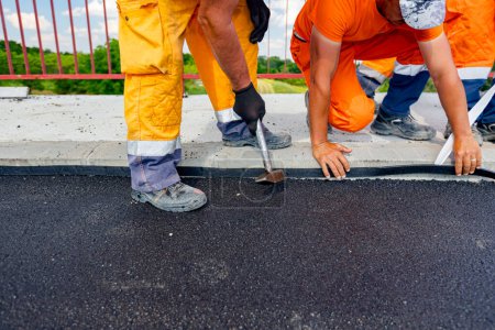Photo for Workers attach a bitumen black tape to protect the roadside of bridge from water. Waterproofing material designed to resist and prevent water from passing through the joint placed on the pavement - Royalty Free Image