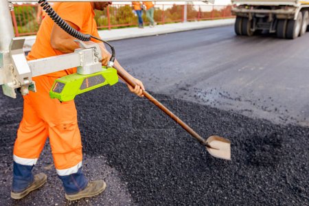 Photo for Ultrasonic grade, slope, non-contact sensor, (multiplex) sensing positioning attached on the road spreading machine, correcting the measured value, height of fresh asphalt, accordingly. Worker uses shovel to level fresh tarmac to right measures. - Royalty Free Image