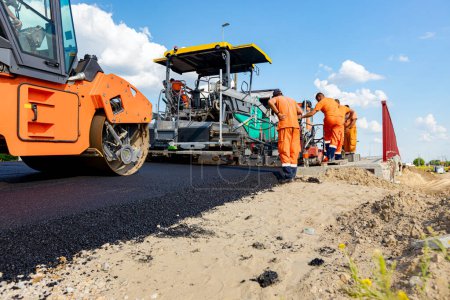 Photo for Hot asphalt is spreading with steamroller. Machine for laying asphalt is spreading layer of hot tarmac on prepared ground a few workers are working around. - Royalty Free Image