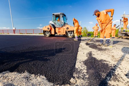 Photo for Zrenjanin, Vojvodina, Serbia - June 8, 2021: Workers are using rakes to level, set up layer of fresh tarmac to right measures, steamroller is flatting fresh asphalt . - Royalty Free Image