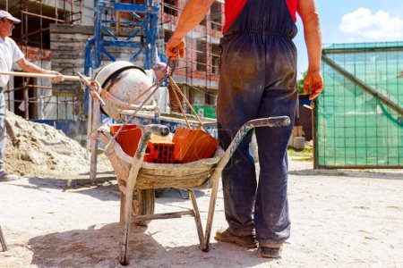 Photo for Construction worker is attaching crane hooks to industrial tipping barrow full of red bricks in the background is mortar mixer machine and edifice under construction with scaffold. - Royalty Free Image