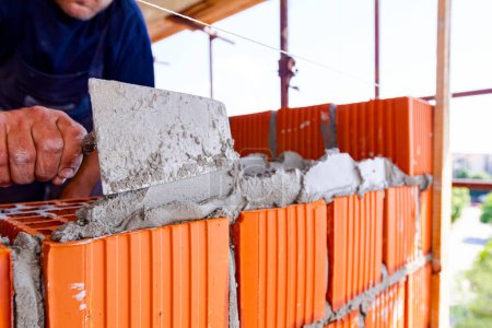 Foto de El trabajador está utilizando espátula, llana, para aplicar mortero en bloques rojos para hacer la pared, albañil. El edificio está en construcción. - Imagen libre de derechos