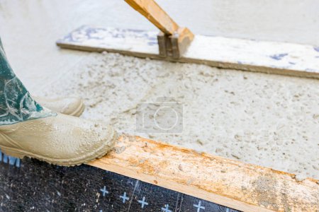 Photo for Rigger in rubber boots, worker is using wooden handmade handcraft tool to leveling fresh concrete after pouring in building foundation. - Royalty Free Image