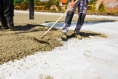 Photo for Construction worker, rigger is using rake to spreading, leveling concrete covering square trench, pouring layer of concrete in foundation. - Royalty Free Image
