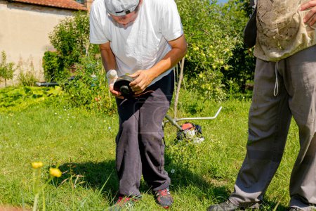 Repairman with bandaged hurt hand repairs trimmer, adjusts string of trimmer head coil, help his friend beekeeper in protective overall to mow grass in orchard and apiary. Maintenance of garden tools